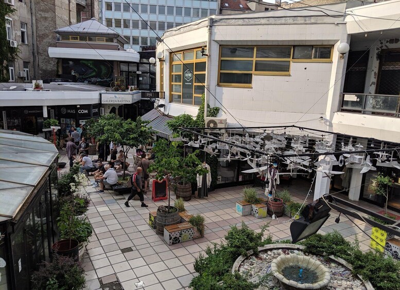 An outdoor market with people walking, stalls selling plants, and a building with a sign 'GALERJA BASTINA'. 2