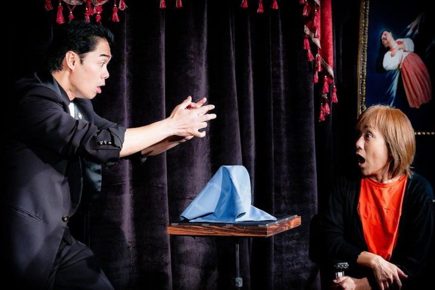 A person in a black suit gesturing towards a blue object on a stand, with a curtain and a framed picture in the background. 3