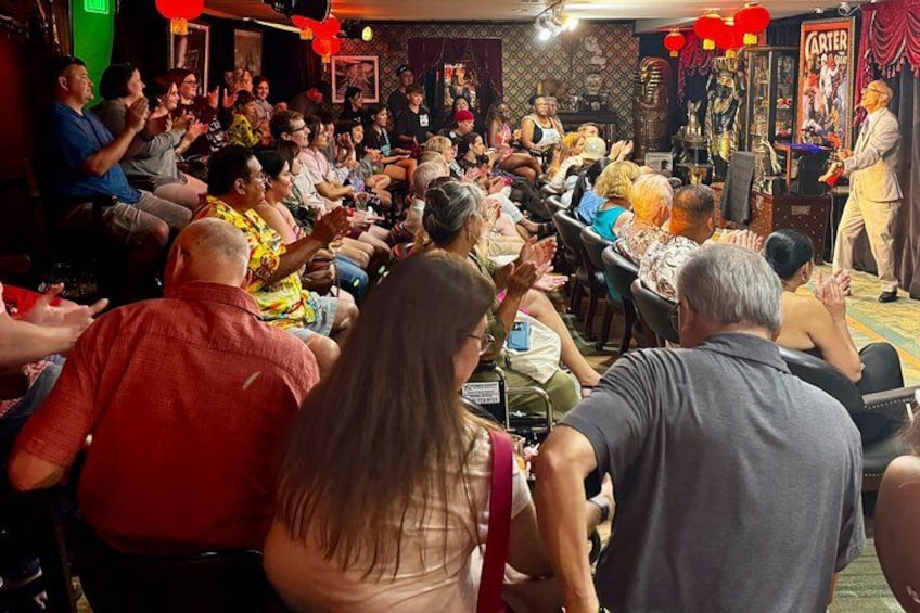 A crowded indoor event with people seated and standing, a stage with a performer, and a poster in the background. 3