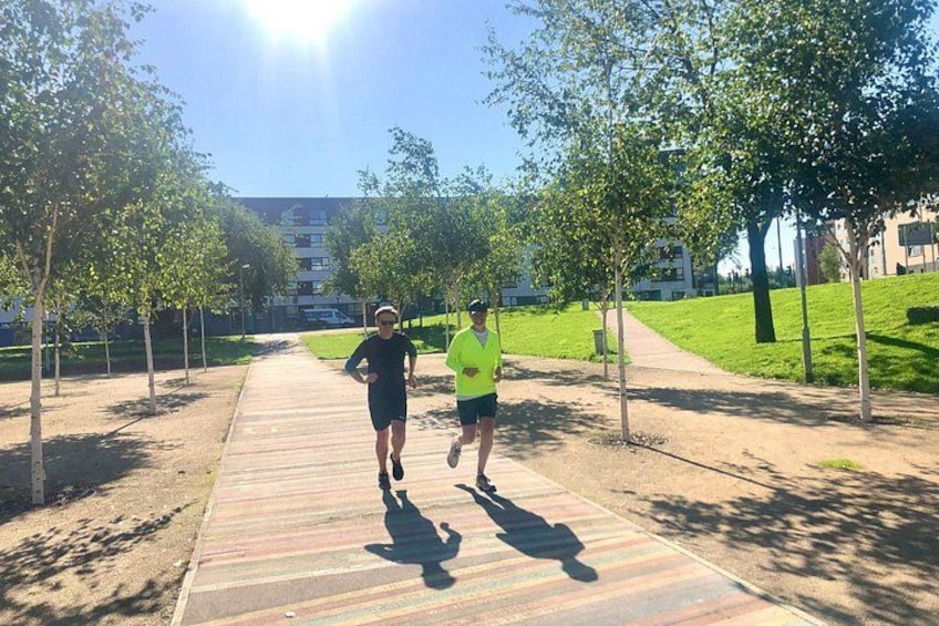 Two people jogging on a paved path surrounded by trees and grass, with buildings in the background. 2