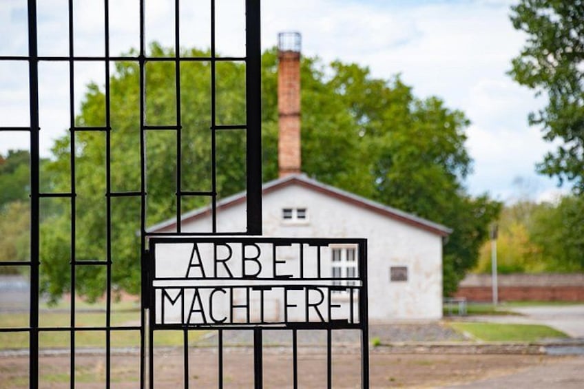 A white building with a chimney, labeled 'ARBEIT MACHTFREI' in front of a metal gate. 2