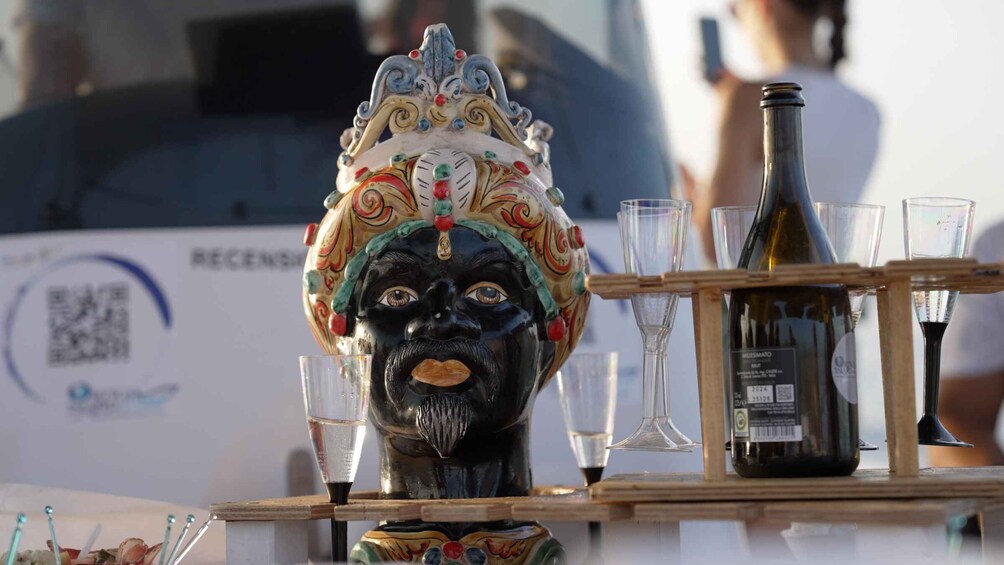 Une tête en céramique décorative, ornée d’un couvre-chef aux couleurs vives, une bouteille de vin et plusieurs verres disposés sur un plateau en bois. 5
