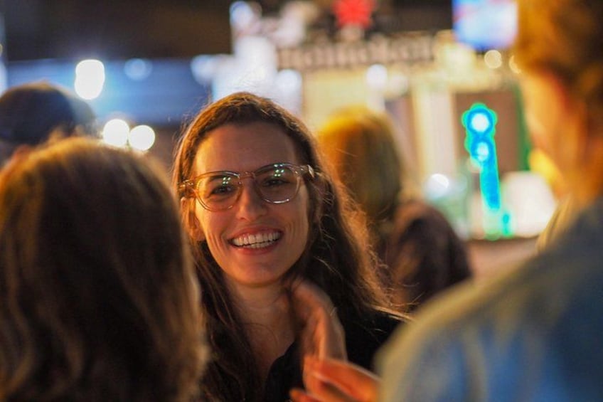 A person with long hair and glasses, wearing a dark top, in a crowded indoor setting with blurred lights in the background. 5