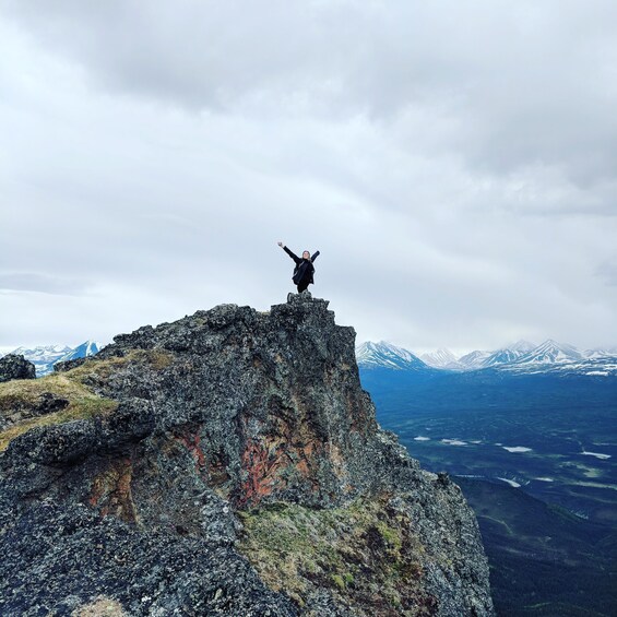 A person standing on a rocky peak with arms raised, overlooking a valley with mountains in the distance. 5