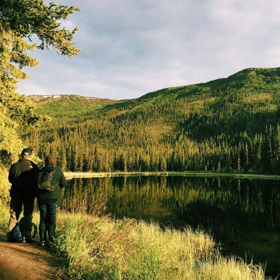 A lake surrounded by forested hills, with two hikers on a trail. 4