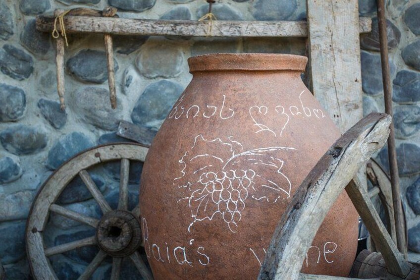 Ein Tonkrug mit Weinranken-Zeichnungen und Schrift, ein hölzernes Rad und eine Mauer aus Stein im Hintergrund. 4