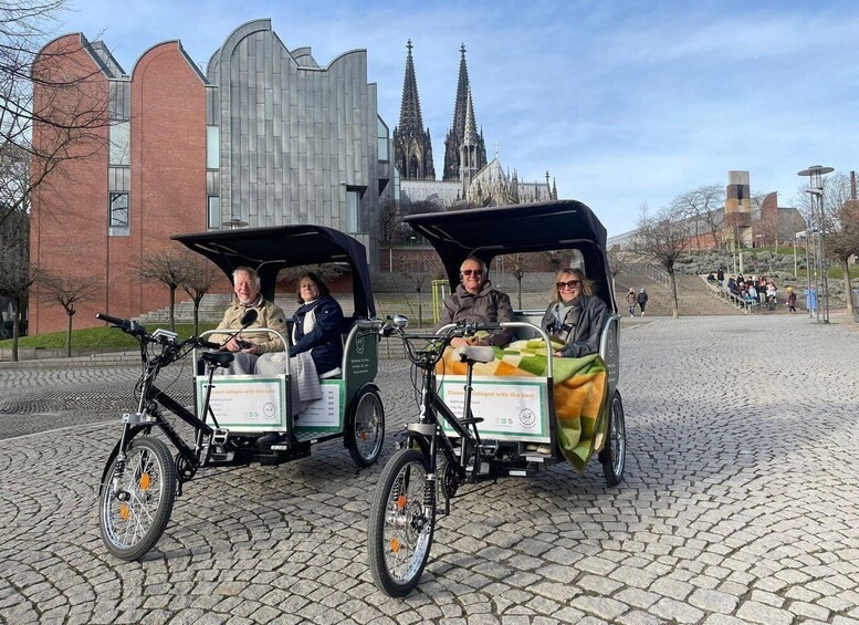 Two people in a covered bicycle carriage with a blanket, parked on cobblestone pavement. 1
