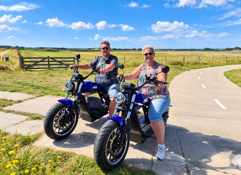 Two people on a blue electric scooter on a paved path with grassy fields and a fence in the background. 2