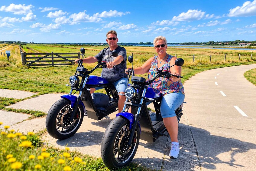 Two people on a blue electric scooter on a paved path with grass and fields in the background. 1