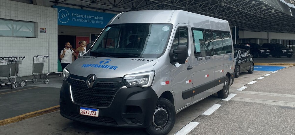 En sølvfarvet varevogn med 'TOP TRANSFER' og 'RJ 935.007' på forsiden, parkeret ved en lufthavnsterminal. 3