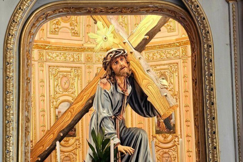 A statue of a figure carrying a cross, set against a detailed architectural backdrop with ornate patterns and a golden frame. 4
