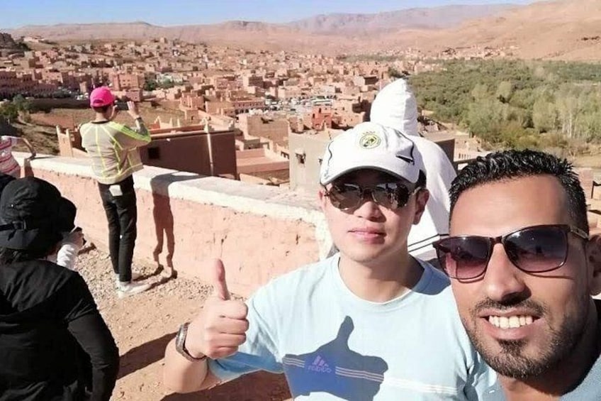 A person giving a thumbs-up gesture, another person taking a photo, and a view of a town with buildings and mountains in the background. 2