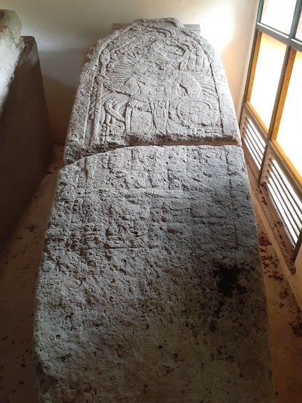 A stone sarcophagus with intricate carvings, placed indoors with a window in the background. 2