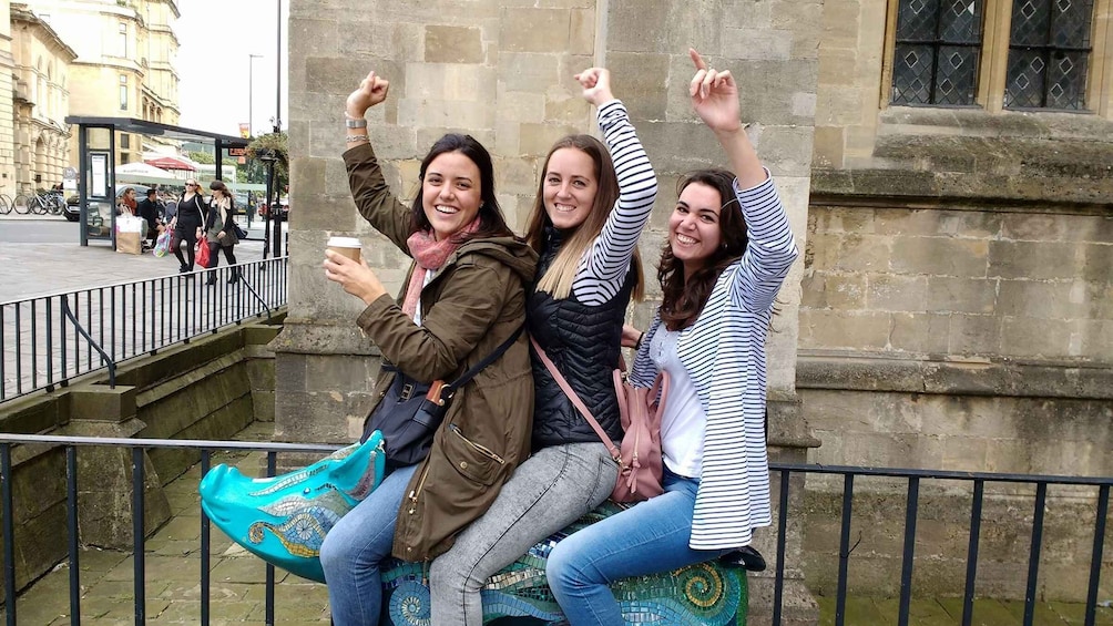 Three women sitting on a bench with raised arms, holding coffee cups. 2