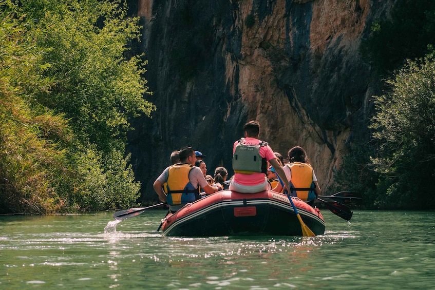 A group of people in a raft navigating a river with a rocky cliff in the background. 1