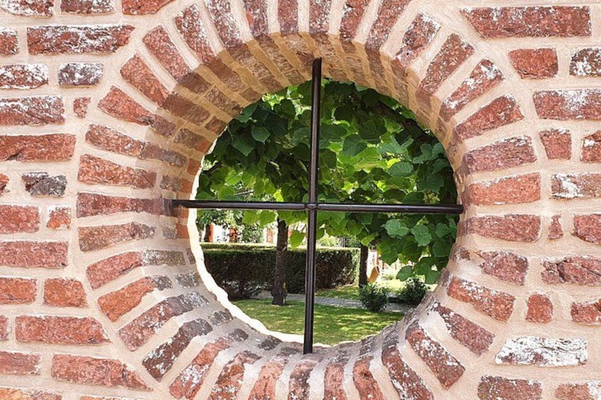 A brick archway with a circular window, a metal cross, and a view of a garden with trees and a hedge. 3