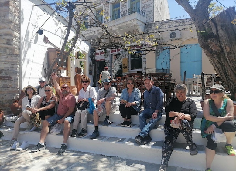 Eine Gruppe von Personen sitzt auf Stufen in einem sonnigen Außenbereich, im Hintergrund sind Gebäude und Bäume zu sehen. 1