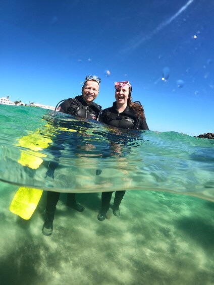 Dos personas con trajes de neopreno de pie en aguas poco profundas con una tabla de surf. 2