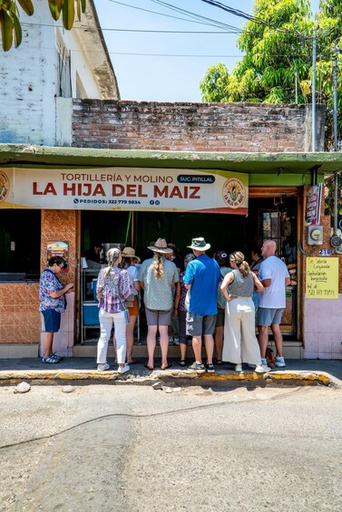 Escena de calle con un grupo de personas paradas frente a una tienda llamada 'La Hija del Maiz'. 5