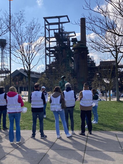A group of people in white vests standing on a sidewalk, facing a large industrial structure with a metal framework and a chimney. 2