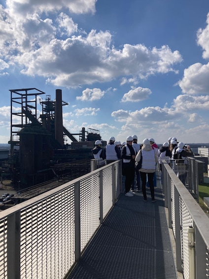 A group of people on a bridge observing an industrial facility with a large chimney. 3