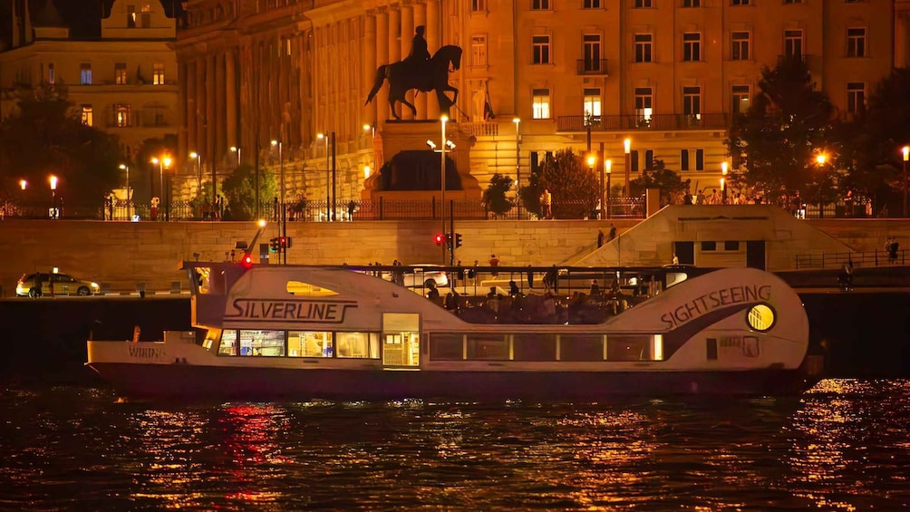 A river cruise boat named 'SIGHTSEEING' with 'SILVERLINE' branding, docked at a waterfront with a statue and illuminated buildings in the background. 5