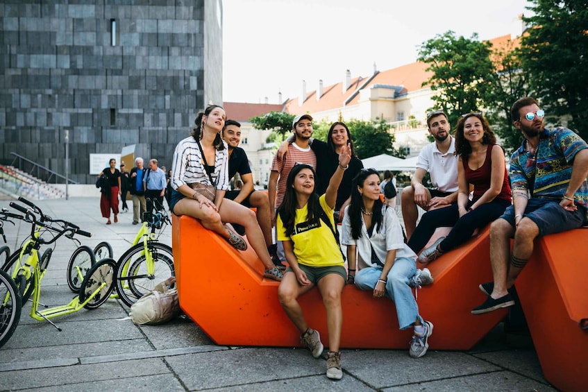 A group of people sitting on orange benches in an urban setting with bicycles and buildings in the background. 1