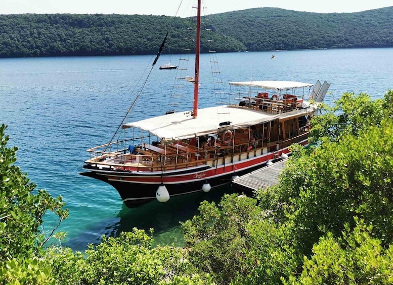 A sailboat docked at a pier with lush greenery in the foreground and a hilly landscape in the background. 2