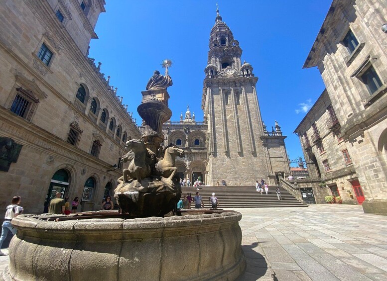 A historic square with a prominent church tower, a fountain with statues, and people walking around. 3