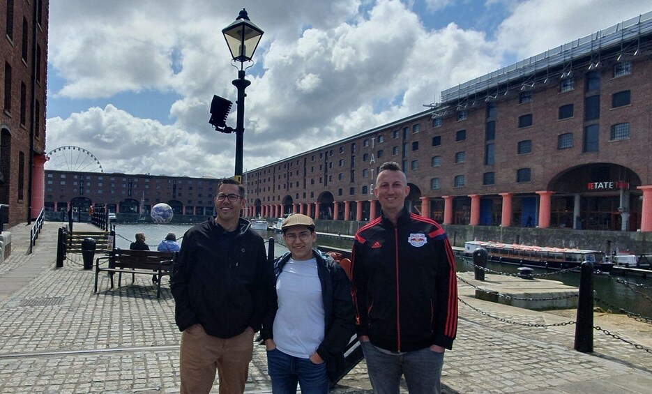 Three people standing on a cobblestone waterfront with a brick building and a Ferris wheel in the background. 1