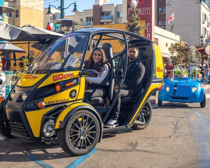 A yellow and black GoCar with two passengers, parked on a street with buildings and a blue car in the background. 2