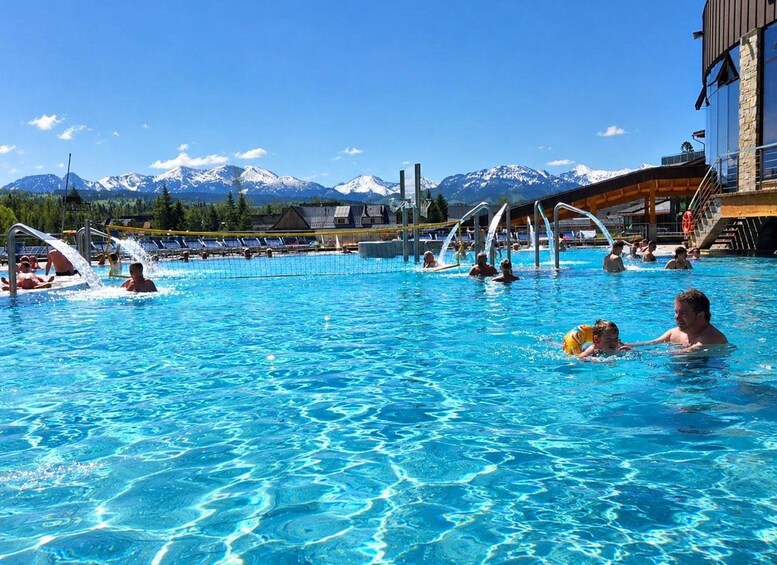 Une piscine où des personnes se baignent et s’amusent, un filet de volley-ball et, en arrière-plan, un paysage montagneux. 1