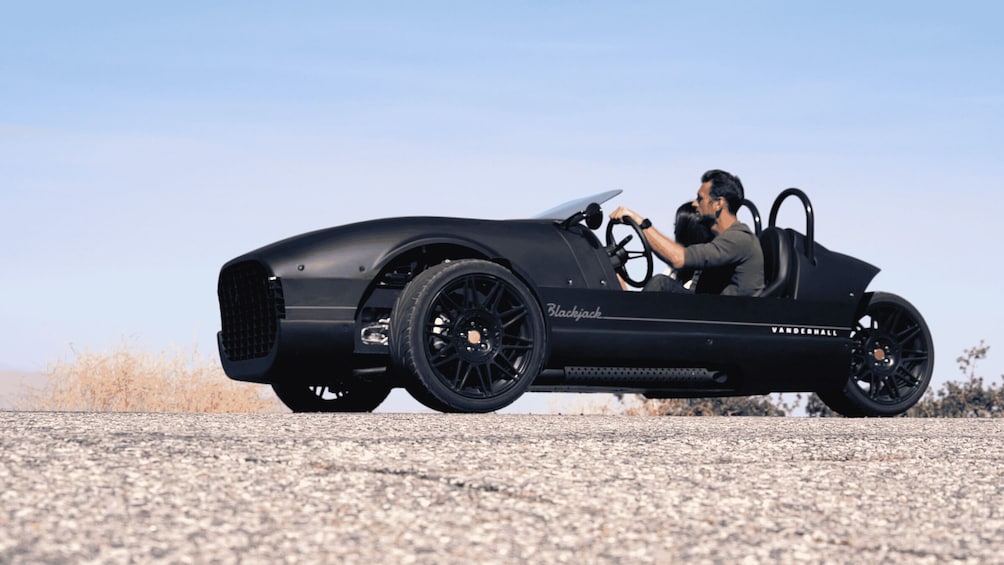 A man driving a vintage car with 'Blackjack' and 'Vanderhall' written on it. 5