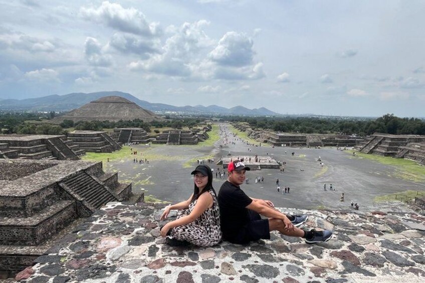 Two people sitting on stone steps overlooking ancient ruins with a large pyramid in the background. 2