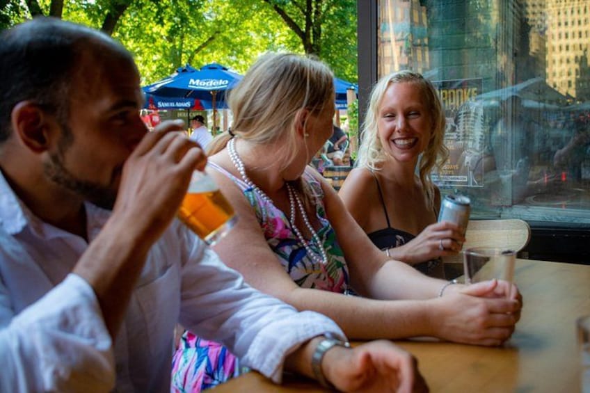 Menschen sitzen draußen an einem Tisch, trinken Bier, mit Sonnenschirmen und einem Gebäude im Hintergrund. 3