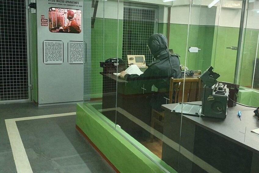 Una persona con sudadera trabajando en un escritorio con una computadora y papeles, en una habitación con paredes verdes y una división de vidrio. 5