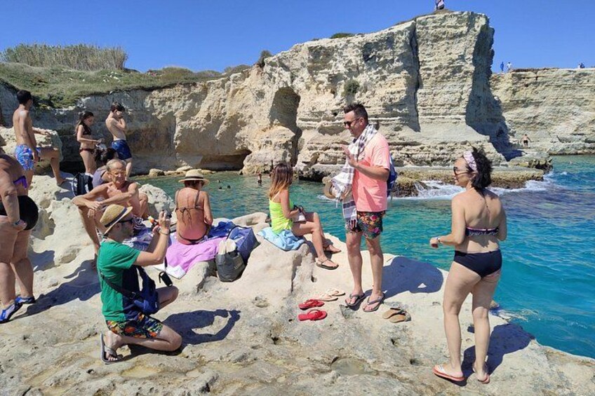 Personas relajándose en una playa rocosa con aguas azules cristalinas y acantilados al fondo. 1