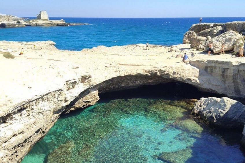 Formación rocosa costera con una piscina de agua cristalina, un arco natural rocoso y un edificio a lo lejos. 5
