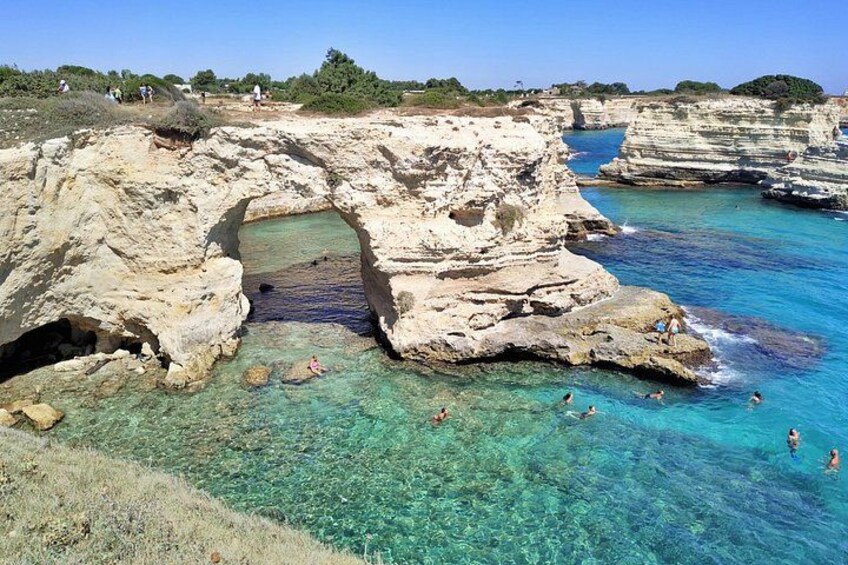 Costa rocosa con agua turquesa transparente, una entrada a cueva y gente nadando. 3