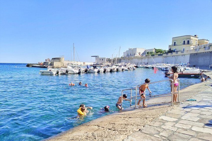 Une scène côtière où l’on voit des gens se baigner et se tenir près de l’eau, des bateaux amarrés et des bâtiments le long de la côte. 5