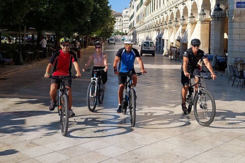 Tres ciclistas pedaleando por un sendero pavimentado, con edificios y árboles al fondo. 3