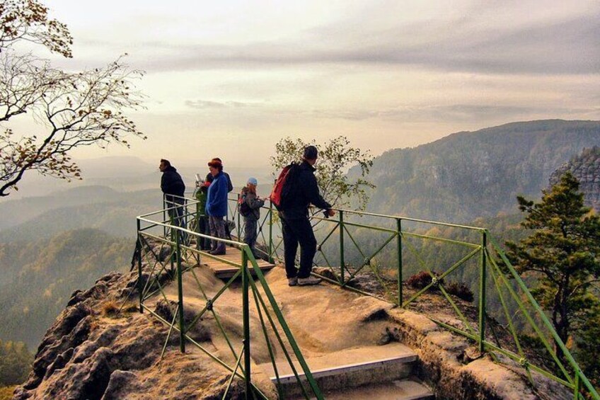 Un grupo de personas de pie sobre un afloramiento rocoso con baranda metálica, contemplando un paisaje montañoso. 4