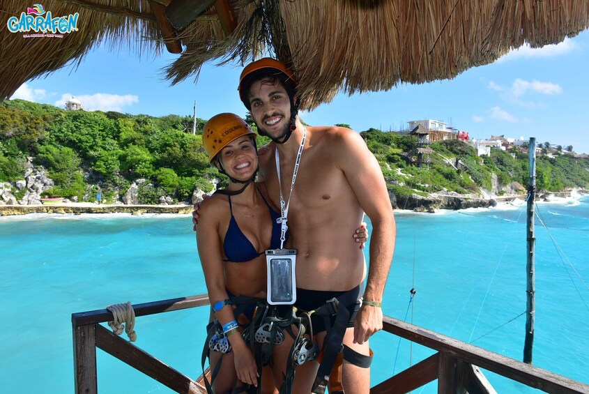 Two people on a wooden platform with a rope harness, overlooking a turquoise beach and a green hillside with buildings. 1