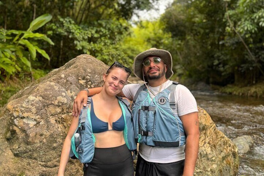 Dos personas caminando por un área boscosa con una gran roca y un arroyo al fondo. 2
