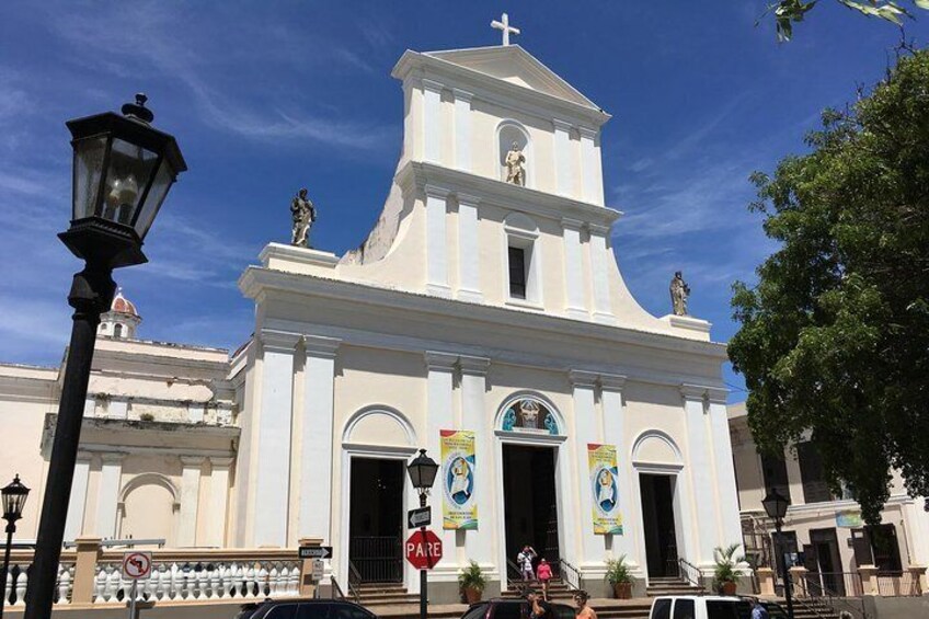 Une église blanche, dotée d’un clocher et de statues sur le toit. Des bannières sont accrochées à l’entrée et un lampadaire se trouve devant. 3