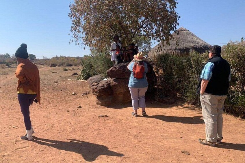 A group of people walking on a dirt path in a dry, open landscape with sparse vegetation and a thatched-roof structure in the background. 2