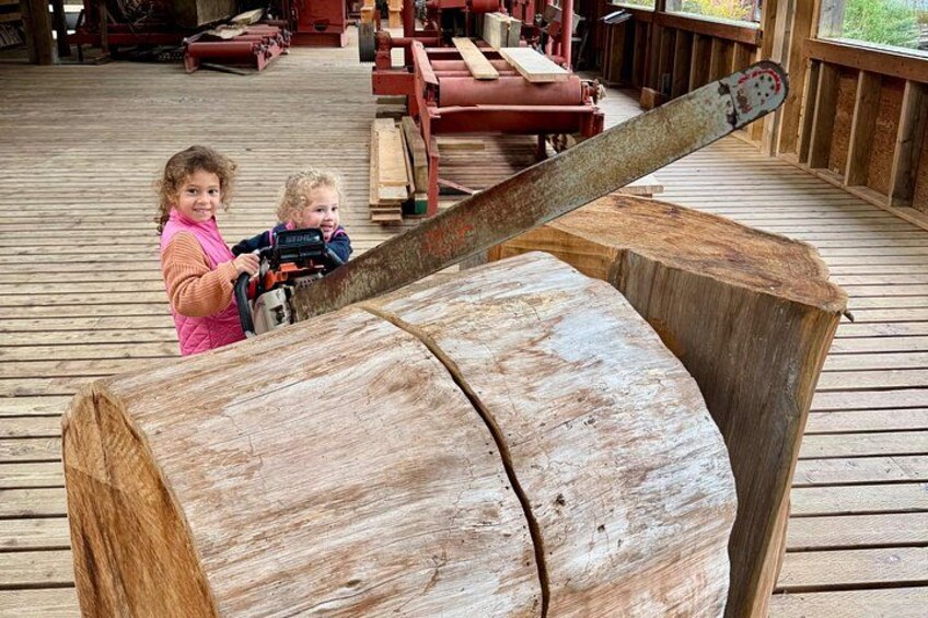Two children on a wooden deck with a large log and a chainsaw. 2