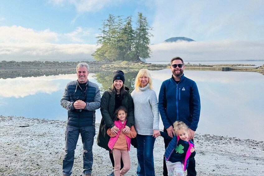 A family of five standing on a sandy beach with a calm lake and a mountain in the background. 1
