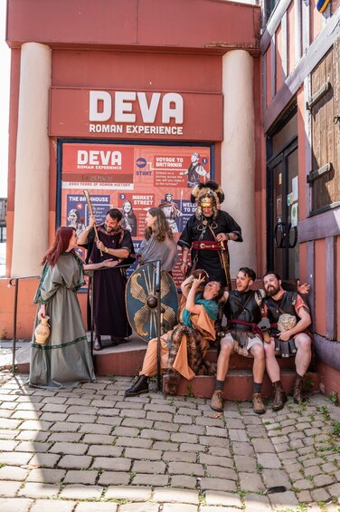 A group of people in Roman attire sitting and standing outside a building with a sign reading 'DEVA ROMAN EXPERIENCE'. 3