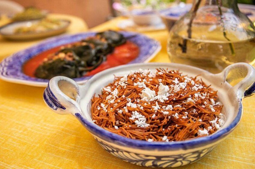 A bowl of noodles garnished with white cheese, a plate of stuffed peppers, and a teapot on a yellow tablecloth. 2